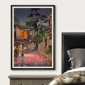 May include: A vintage Japanese woodblock print depicting a rainy night scene in a narrow street lined with traditional houses. A woman with an umbrella walks down the street, illuminated by lanterns. The print is framed in black.