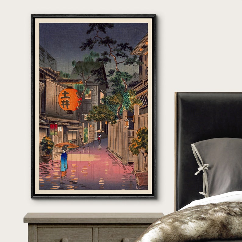May include: A vintage Japanese woodblock print depicting a rainy night scene in a narrow street lined with traditional houses. A woman with an umbrella walks down the street, illuminated by lanterns. The print is framed in black.