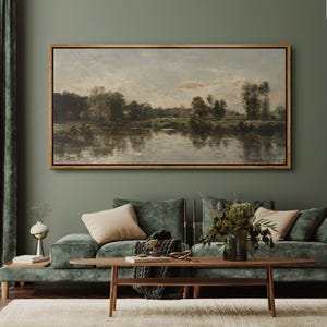 May include: A framed landscape painting depicting a river scene with trees and a cloudy sky. The artwork is in a wooden frame and hangs above a green velvet sofa with beige pillows. A wooden coffee table and decorative items complete the room.