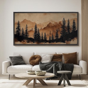 May include: A framed landscape art print featuring a watercolor painting of a forest and mountains in shades of brown and black. The artwork hangs above a white sofa with decorative pillows and a wooden coffee table.