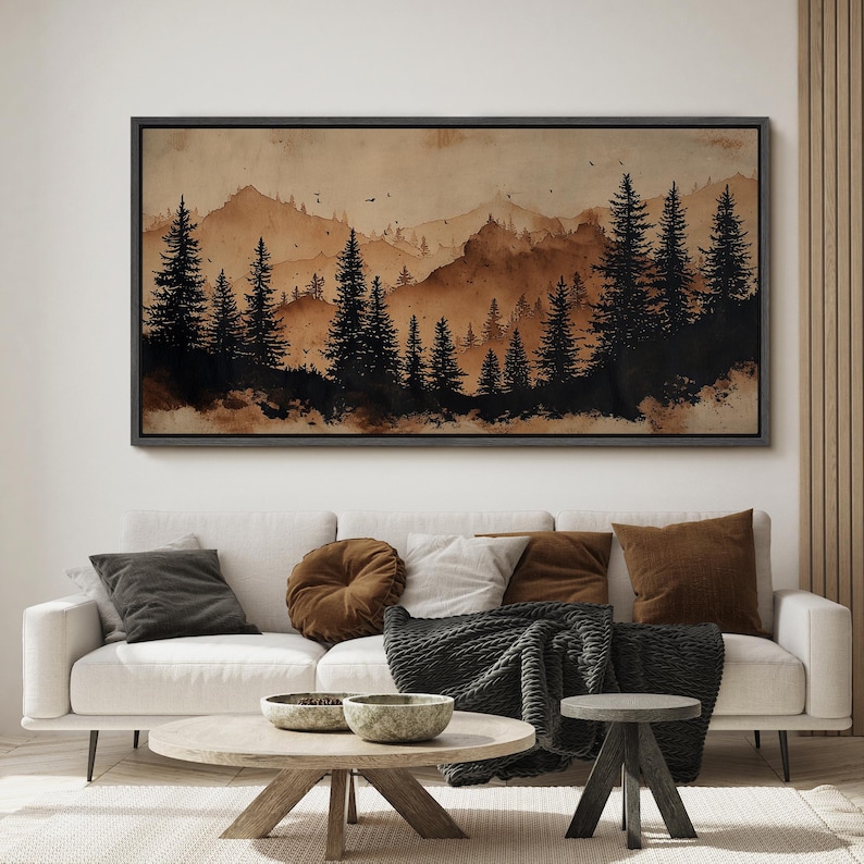 May include: A framed landscape art print featuring a watercolor painting of a forest and mountains in shades of brown and black. The artwork hangs above a white sofa with decorative pillows and a wooden coffee table.