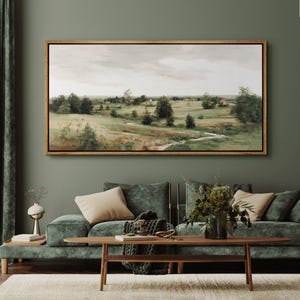 May include: A framed landscape painting with a wooden frame hangs above a green velvet sofa. The painting depicts a serene countryside scene with rolling hills, trees, and a cloudy sky. The room is decorated with a coffee table, throw pillows, and decorative items.