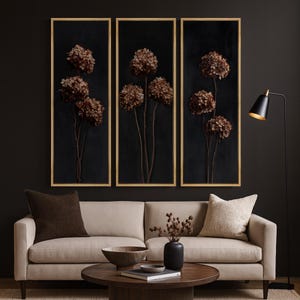 May include: Three framed wall art pieces featuring dried brown hydrangeas against a black background. The frames have a gold finish. The artwork is displayed above a beige sofa with brown and beige accent pillows. A black floor lamp and a round coffee table complete the room.