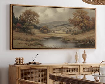 May include: A framed landscape painting depicts a serene river scene with trees in autumn colors. The artwork features a wooden frame and is displayed above a wooden cabinet with decorative items.