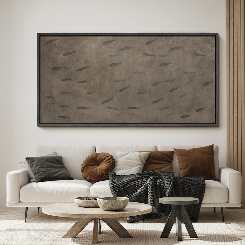 Puede incluir: Una gran obra de arte en lienzo enmarcada rectangular que presenta un banco de peces nadando en un fondo de tonos marrones. La obra de arte cuelga sobre un sof&aacute; blanco con varias almohadas y una manta. Una mesa de centro y una mesa auxiliar de madera est&aacute;n al frente.