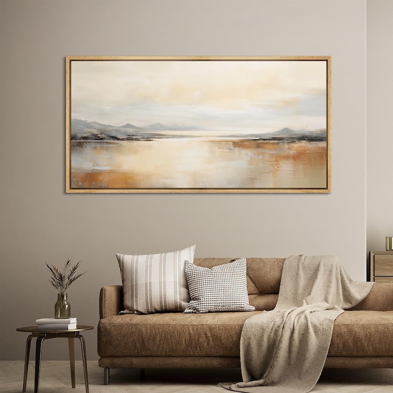 May include: A horizontal abstract landscape painting with a muted color palette of beige, brown, and gray. The painting depicts a lake with mountains in the distance. The painting is framed in a light wood frame.