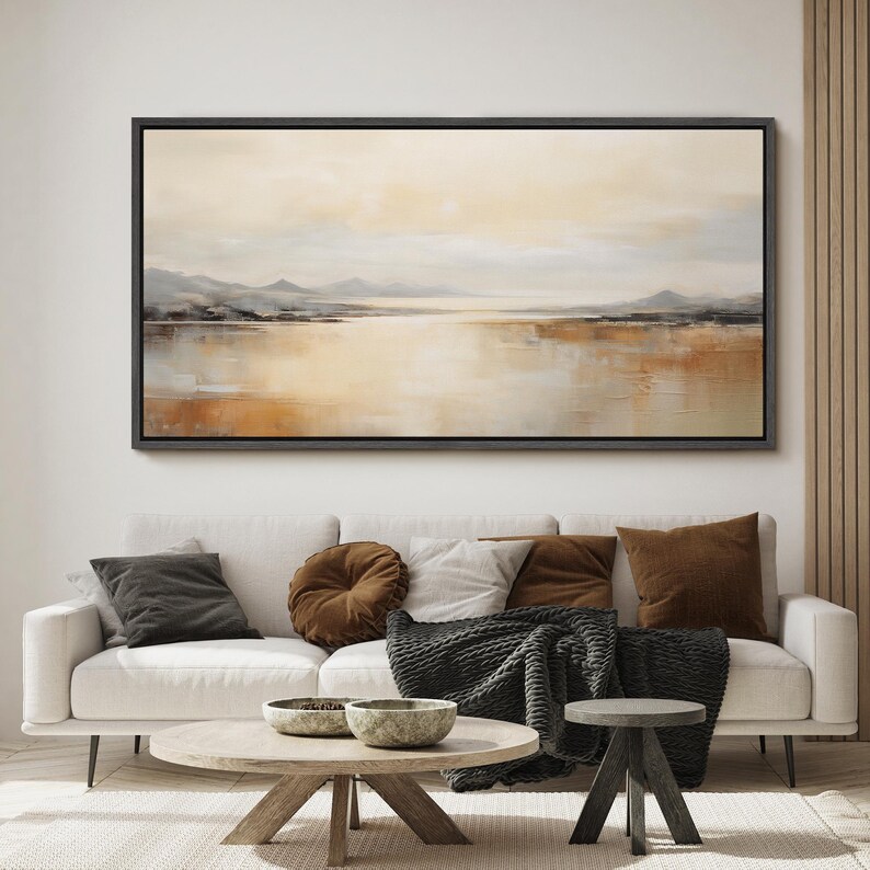 May include: A large abstract landscape painting in muted tones of brown, beige, and gray. The painting depicts a lake or river with mountains in the distance. The painting is framed in a black frame and is hanging on a white wall.