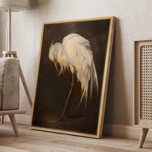 May include: A framed artwork shows a white egret with its head lowered, set against a dark brown backdrop. The bird's feathers are detailed with soft, light colours. The frame is made of light-coloured wood.