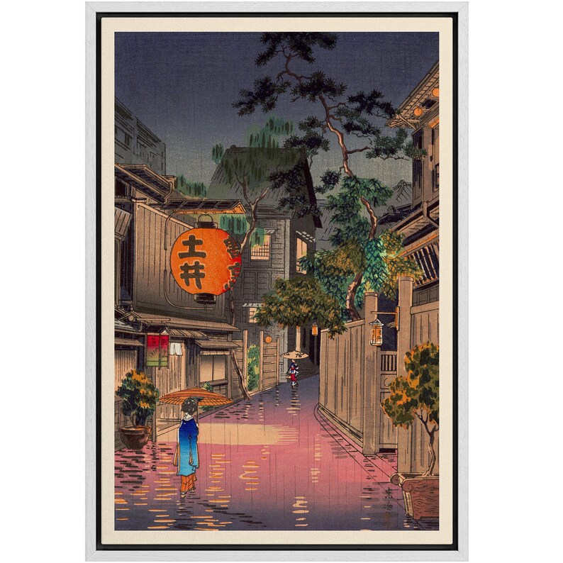 May include: A woodblock print depicting a narrow street in a Japanese city at night. The street is wet from rain and lit by a single lantern hanging from a building. A woman in a blue kimono walks down the street, carrying an umbrella. The print is framed in a white frame.