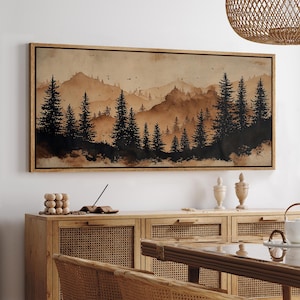 May include: A framed landscape art print featuring a watercolor-style forest scene in shades of brown and black. The artwork depicts silhouetted pine trees in the foreground with mountains in the background. The frame is made of light-colored wood.