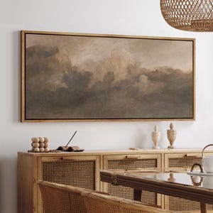 May include: A framed landscape painting depicts a dramatic sky filled with dark, swirling clouds in shades of brown and gray. The artwork is displayed above a wooden cabinet with woven cane doors, complemented by decorative items.
