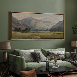 May include: A framed landscape painting depicts a mountain range with a valley and a field of green grass. The artwork has a wooden frame and is displayed above a green sofa. The color palette includes shades of green, gray, and brown.