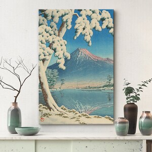 Puede incluir: Una estampa en madera que representa el monte Fuji en la distancia, con un &aacute;rbol cubierto de nieve en primer plano y un lago que refleja la monta&ntilde;a. El cielo es azul y el agua es azul claro.