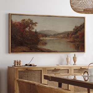 May include: A framed landscape painting depicts a serene river scene with autumn foliage. The artwork features a wooden frame and showcases a palette of warm colors, including reds, oranges, and browns, reflecting the season. The painting evokes a sense of tranquility.