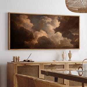 May include: A framed rectangular artwork depicts a dramatic cloudscape in shades of brown, beige, and white. The artwork is mounted in a light brown wooden frame and hangs above a wooden cabinet. The overall aesthetic is warm and inviting.