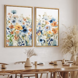 May include: Two framed watercolor paintings of blue, orange, and yellow flowers on a white background. The paintings are in gold frames and are hanging on a white wall.