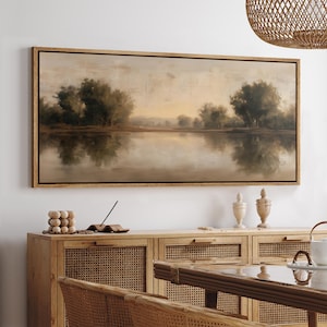 May include: A rectangular landscape painting with a wooden frame. The artwork depicts a serene lake scene with trees reflected in the water. The color palette is muted, featuring shades of beige, brown, and green, creating a calming atmosphere. The painting is displayed above a wooden cabinet.