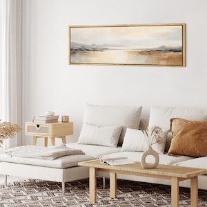 May include: A living room interior with a white couch, a wooden coffee table, and a framed landscape painting above the couch. The painting features a muted color palette of blues, browns, and grays, depicting a lake with mountains in the distance.