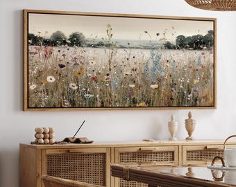 May include: A framed landscape painting depicts a field of wildflowers in a meadow. The artwork features a variety of colorful blooms, including white, yellow, red, and blue flowers, set against a soft, neutral background. The painting is encased in a light brown wooden frame.