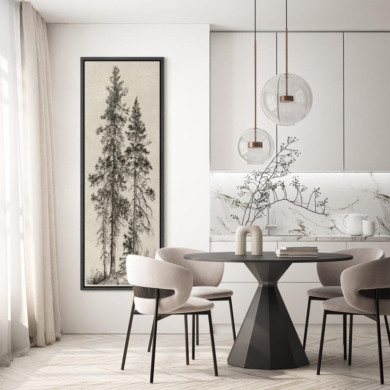 May include: A framed black and white print of two tall pine trees. The artwork hangs on a white wall in a modern dining room setting. The room features a round black table, chairs, and pendant lights.