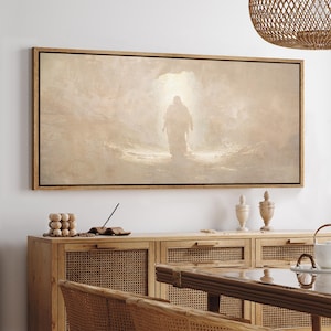 May include: A framed canvas art print depicts a figure walking towards a bright light. The artwork is in muted tones of beige and brown, with a dark wooden frame. The image is mounted on a white wall above a wooden cabinet.