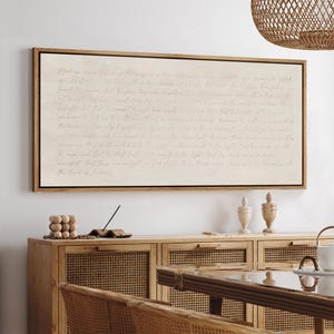 May include: A framed rectangular artwork with handwritten text in a light beige hue. The wooden frame is a warm brown. The artwork is displayed above a wooden cabinet with woven panels, and a dining table with a woven chair.