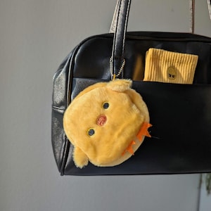 Könnte beinhalten: Eine schwarze Handtasche mit einem gelben Plüsch-Küken-Schlüsselanhänger. Ein gelbes Portemonnaie mit einem Küken-Gesicht befindet sich in der Vordertasche. Der Küken-Schlüsselanhänger hat schwarze Augen und einen orangefarbenen Schnabel. Die Tasche hat einen oberen Griff und einen Schulterriemen.