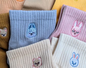 May include: A collection of colorful socks in shades of tan, yellow, pink, and blue. Each sock features an embroidered animal face, including a cat, dog, and bunny. The socks are made of a ribbed knit material.