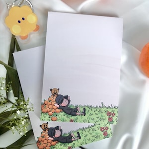 Può includere: Due biglietti vuoti con un'illustrazione di un cartone animato di una persona sdraiata su un mucchio di arance con un piccolo orso. L'illustrazione è circondata da erba verde e fiori rossi.