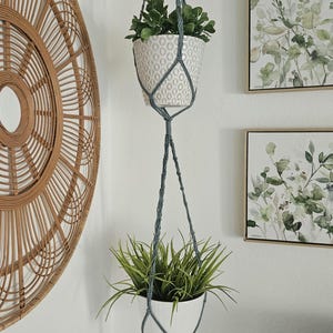 Può includere: Due vasi di ceramica bianca con piante verdi sospesi da un appendiabiti in macramè grigio. Il vaso superiore ha un motivo. Lo sfondo include decorazioni murali tessute e arte botanica incorniciata.