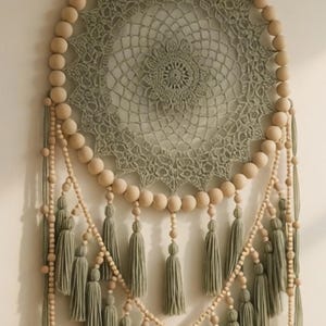 May include: A large, round dream catcher with a sage green crocheted centre and wooden beads. The dream catcher has multiple rows of sage green tassels and is suspended by a natural-coloured cord.