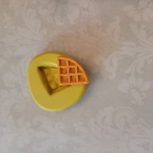 May include: A green silicone mold shaped like a triangle with a waffle pattern inside. A small, brown waffle is sitting in the mold.