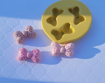 May include: A yellow silicone mold with four bow-shaped cavities. Three pink bows with white polka dots are shown outside of the mold.