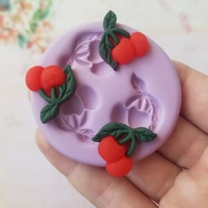 May include: Silicone mold with three impressions of cherries with green leaves. The mold is purple and the cherries are red.