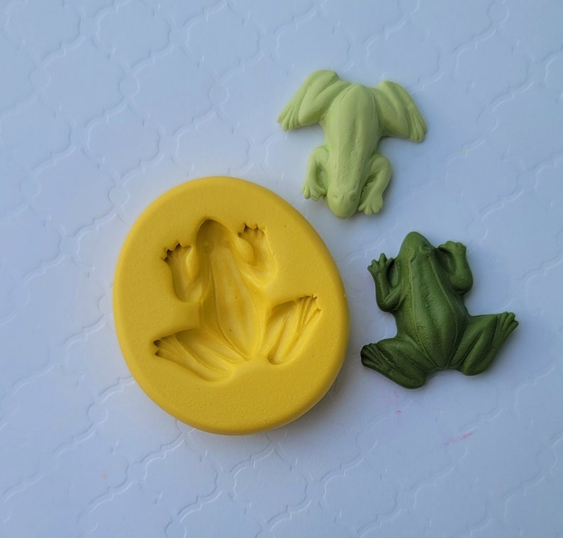 May include: A yellow silicone mould for making frog-shaped decorations. Two green frogs are shown next to the mould.