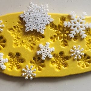 May include: Yellow silicone mold with 10 snowflake designs.  There are 7 white snowflake decorations on top of the mold.