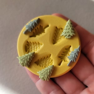 May include: A yellow silicone mold with six impressions of pine trees. Five of the impressions are filled with green and blue polymer clay pine trees.