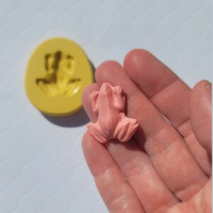 May include: A pink frog figurine made from resin. The frog is sitting with its legs outstretched and its mouth slightly open.