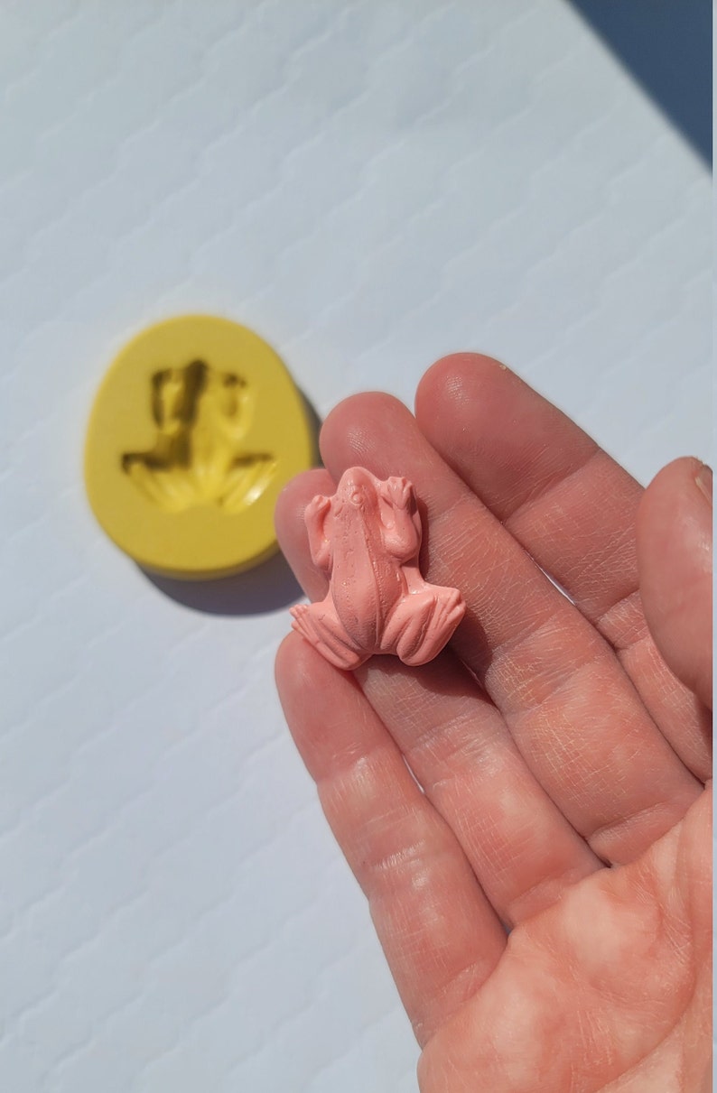 May include: A pink frog figurine made from resin. The frog is sitting with its legs outstretched and its mouth slightly open.