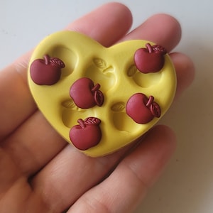 May include: A yellow heart-shaped silicone mold with six individual cavities shaped like red apples. The mold is held in a hand.