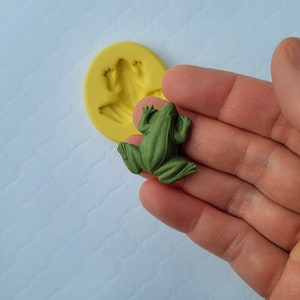 May include: A green frog figurine, made of resin, with a detailed texture. The frog is sitting on a hand, with a silicone mould of a frog in the background.