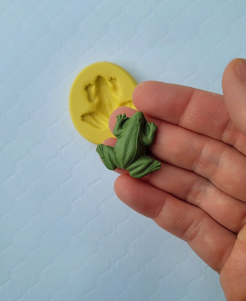 May include: A green frog figurine, made of resin, with a detailed texture. The frog is sitting on a hand, with a silicone mould of a frog in the background.