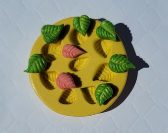 May include: A yellow, circular mold with leaf-shaped cavities. The mold is filled with green, yellow, and pink leaf-shaped decorations. The leaves have detailed veining and are arranged around the edge of the mold.