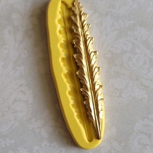 May include: A yellow silicone mold with a gold-colored leaf design. The mold is oval-shaped and features a detailed impression of the leaf. The gold leaf is placed inside the mold, showcasing the design. Ideal for crafting or decorative projects.