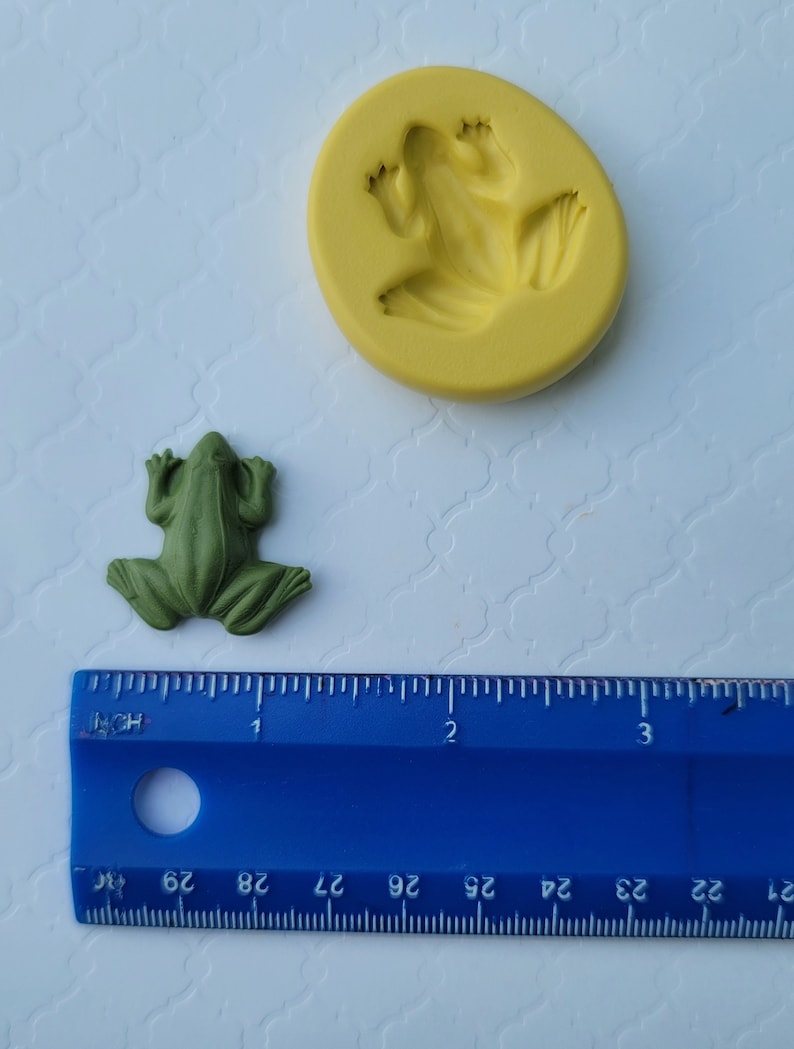 May include: A green frog figurine and a yellow silicone mould of a frog. The mould is about 5 cm in diameter. A blue ruler is in the background.