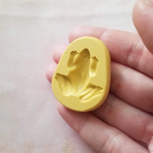 May include: Yellow silicone mould for making a frog-shaped object. The mould is in the shape of a frog with its legs outstretched.