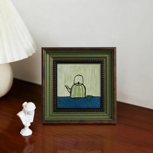 May include: A square framed painting featuring a light green teapot and a small matching green cup on a dark blue surface against a pale green background. The frame is dark green with a textured, beaded inner border and a dark brown outer edge.