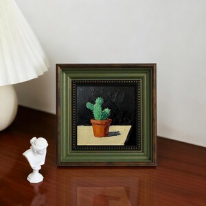 Può includere: Dipinto incorniciato di un cactus in un vaso di terracotta. Il dipinto ha una cornice verde scuro con un bordo interno perlato. Il cactus è verde e il vaso è arancione. Lo sfondo è nero e il tavolo è beige.