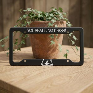 May include: Black license plate frame with the text "YOU SHALL NOT PASS!" in white. The frame has a minimalist design with a stylized illustration. A potted plant with green leaves is in the background.