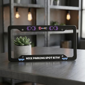 Op de afbeelding: Zwart kentekenplaatraam met de tekst "NICE PARKING SPOT RITA!". Het frame heeft paarse brillen en een halterontwerp. Een kleine vetplant in een betonnen pot staat achter het frame.